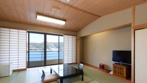 A traditional Japanese room with tatami flooring, a low table with cushions, a television on a wooden cabinet, and a sliding door leading to a balcony with a view of the sea. - Kaisen no Yado Matsuura (Saiki)