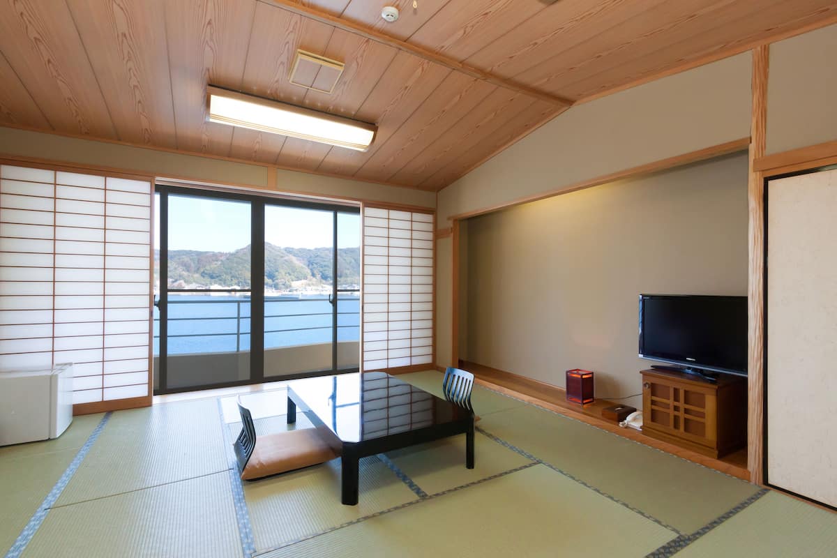 Ein traditioneller japanischer Raum mit Tatami-Böden, einem niedrigen Tisch mit Kissen, einem Fernseher auf einer Holzkommode und einer Schiebetür, die zu einem Balkon mit Meerblick führt.