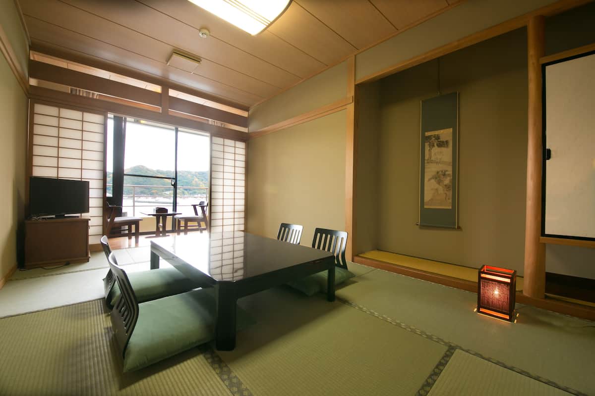 Ein traditioneller japanischer Raum mit Tatami-Boden, einem niedrigen Tisch, Stühlen, einem Fernseher und Blick ins Freie.
