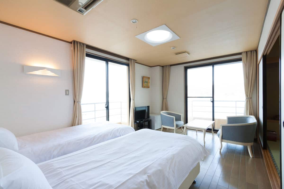 Ein Hotelzimmer mit zwei Betten, einem Schreibtisch, einem Sessel, einem Fernseher und großen Fenstern.