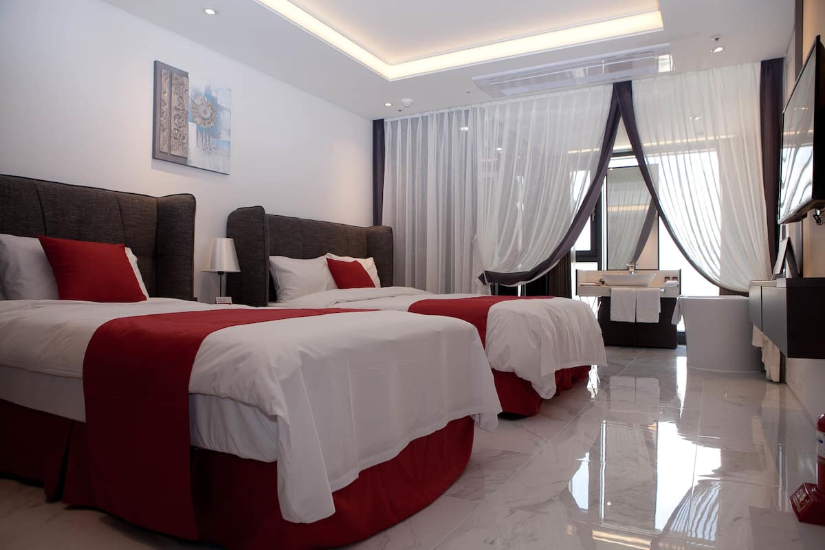Une chambre d’hôtel moderne avec deux lits, une coiffeuse et une grande fenêtre agrémentée de rideaux légers.