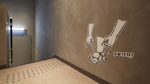 Un pasillo con piso texturizado, una puerta a la izquierda y una pared con una ilustración de un personaje de caricatura y la palabra 'SWISH!'.