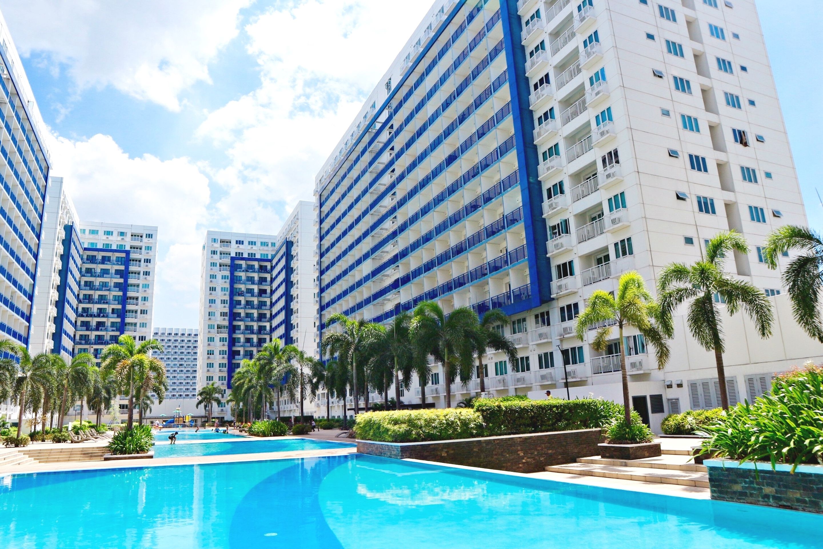 A modern hotel with multiple high-rise buildings, a central swimming pool, and palm trees.