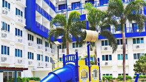 A playground with a blue slide and yellow play structures, set against a backdrop of a white building with blue accents and balconies. - CondoDeal at Sea Residences (Pasay)