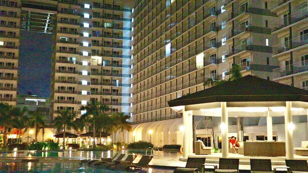 A hotel pool area with lounge chairs, a covered seating area, and a large building in the background. - CondoDeal at Sea Residences (Pasay)