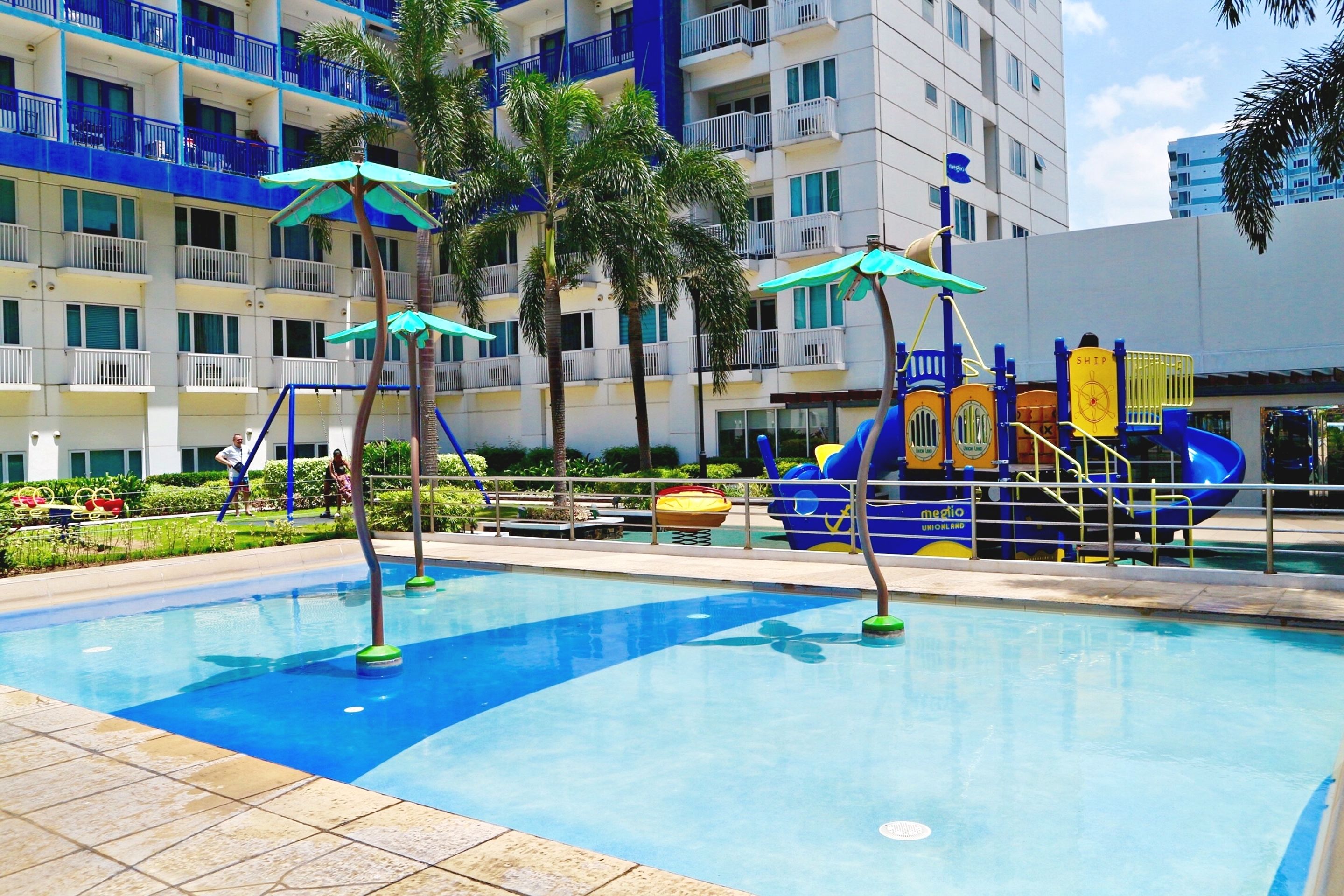 A swimming pool with a children's play area in the background, surrounded by palm trees and a multi-story building with balconies.