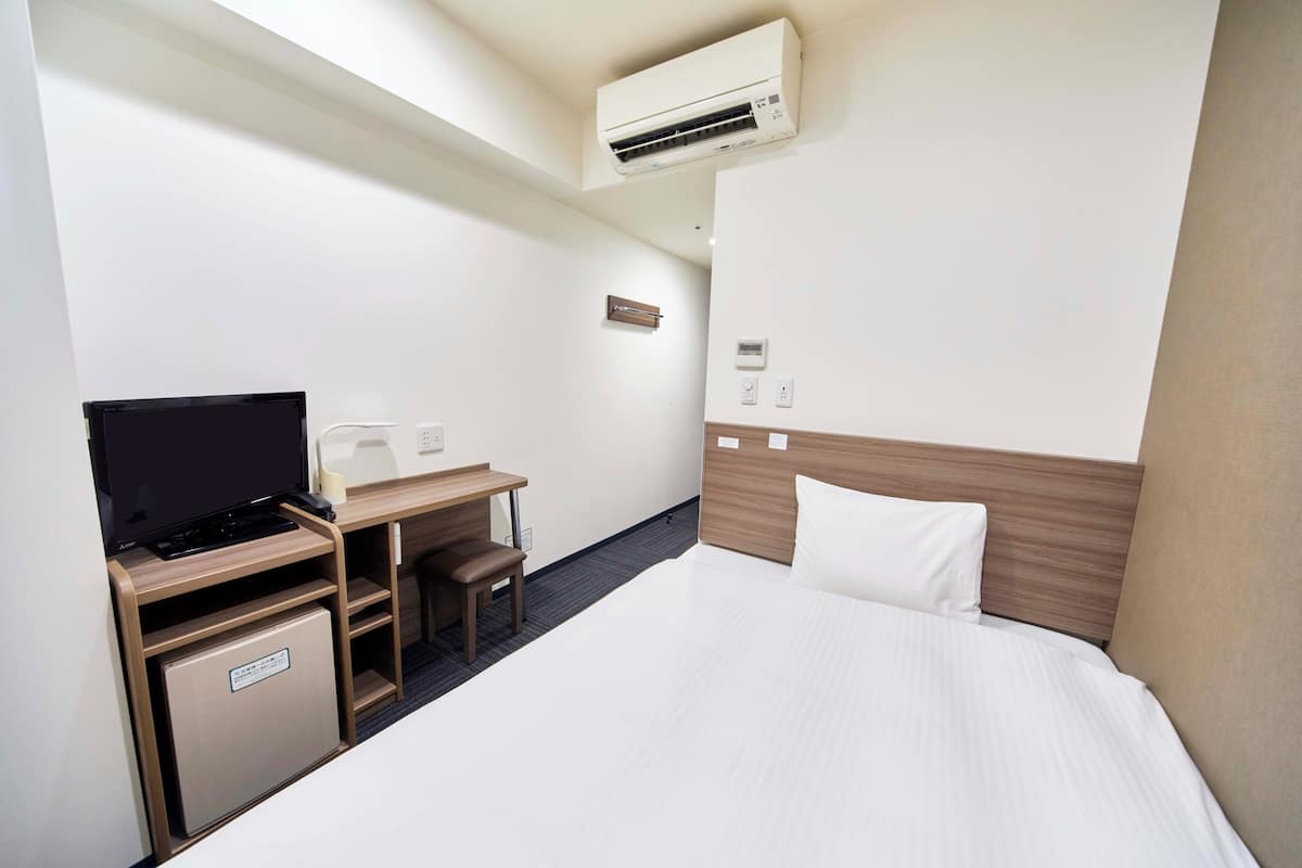 A hotel room with a bed, a desk with a television, a small refrigerator, and an air conditioning unit.