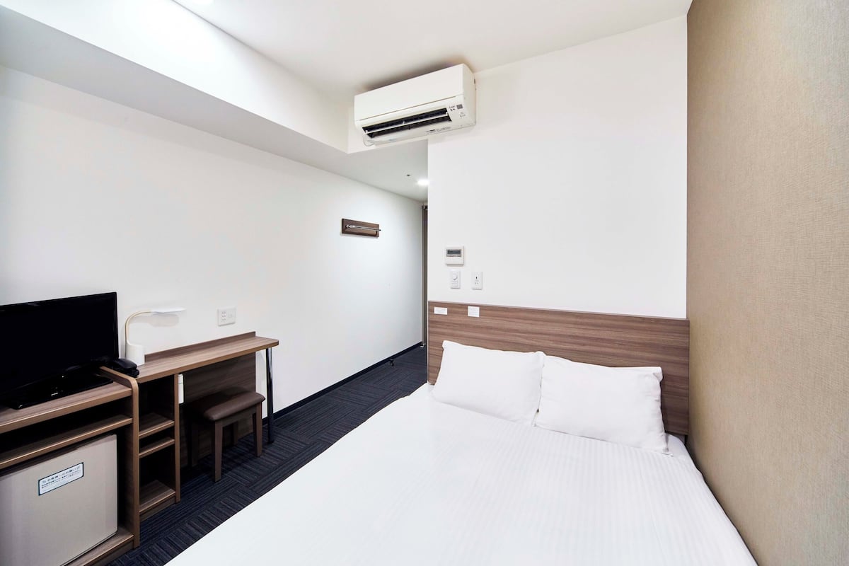 A hotel room with a bed, a desk with a chair, a television, and an air conditioning unit.