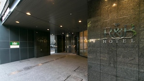 Une entrée d'hôtel moderne, avec une enseigne R&B Hotel bien visible, un panneau vert et un sol carrelé.