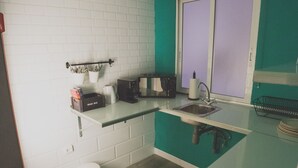 Een kleine keuken met witte bakstenen muren, een blauwgroen aanrecht, een spoelbak, een koffiezetapparaat en een raam met gezandstraalde ruit.