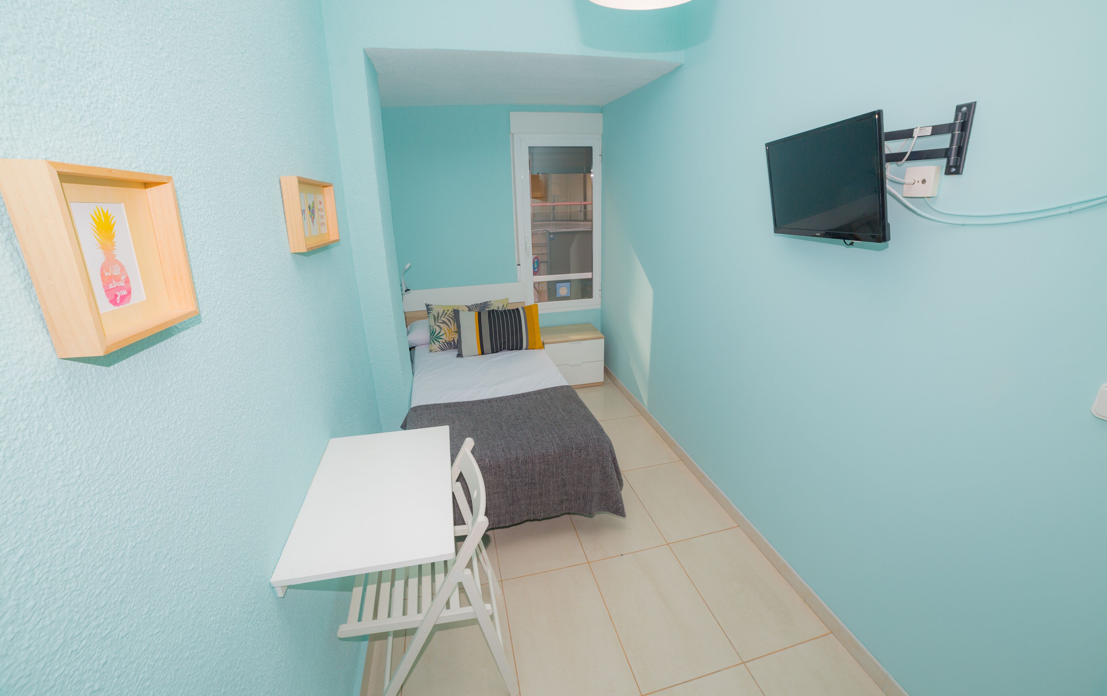 Una habitación pequeña y estrecha con una cama, un escritorio y una televisión instalada en la pared.