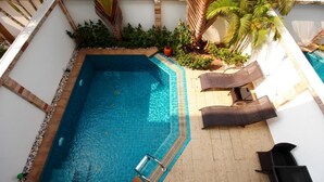 Una piscina rectangular con borde de azulejos, rodeada por un muro blanco y un solárium también con azulejos. Se observan dos sillas de descanso y una pequeña mesa con un objeto decorativo.