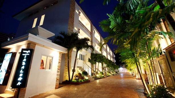 Un hotel moderno con una entrada bien iluminada, una escalera visible y una fila de palmeras a lo largo del sendero.