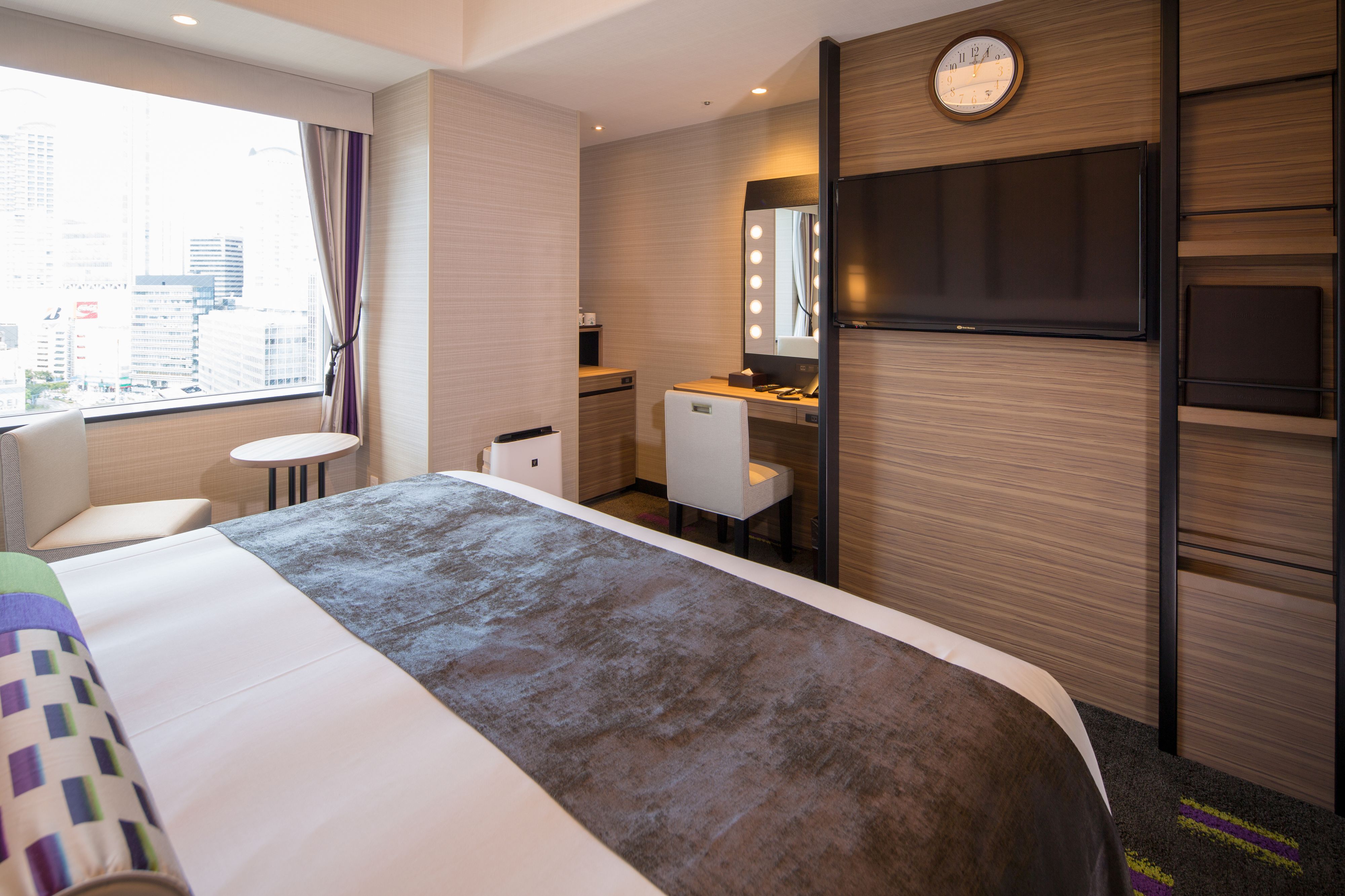A hotel room with a bed, a desk with a mirror and lights, a small table, and a view of the city.