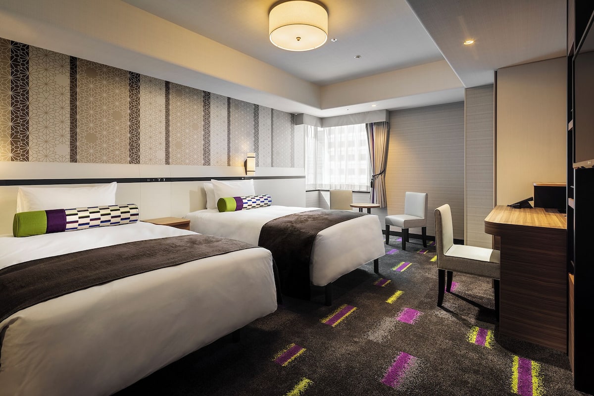 A hotel room with two beds, a desk, a chair, and a large window with curtains.