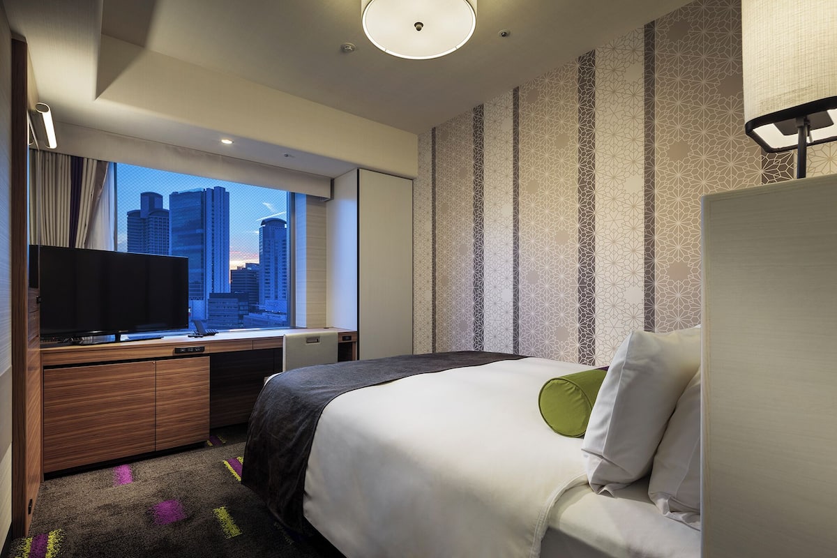A hotel room with a bed, a desk with a television, a window with a city view, and patterned wallpaper.