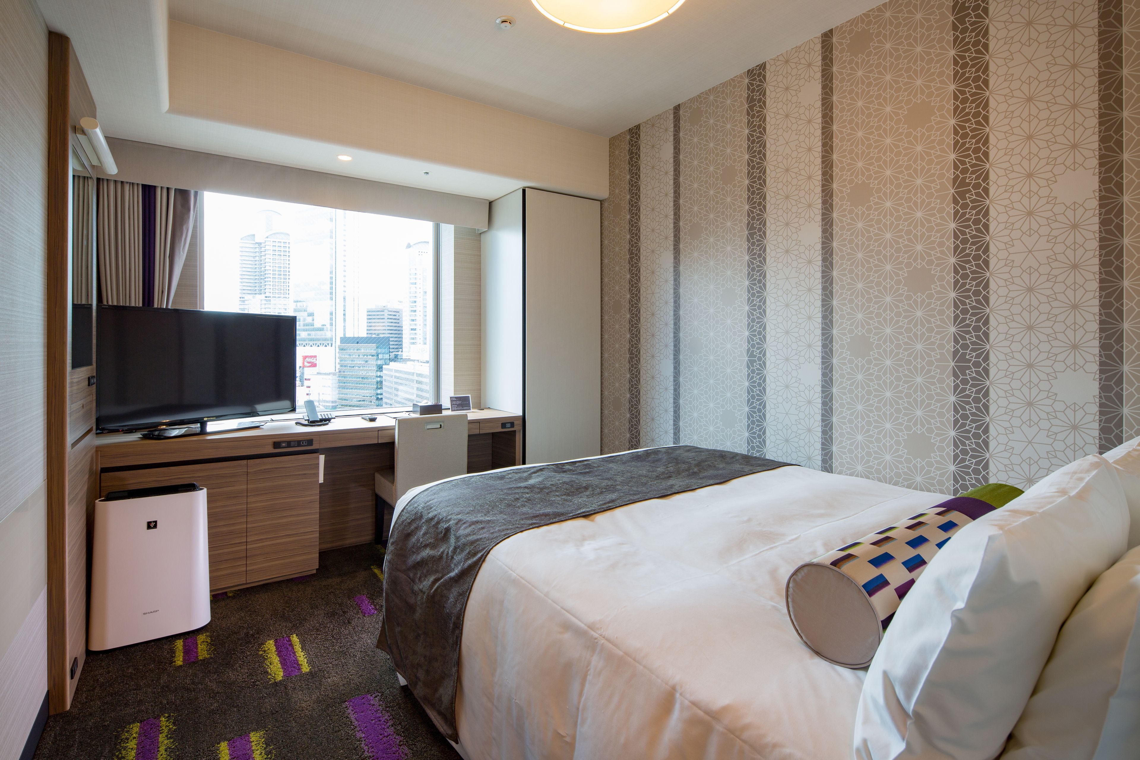 Une chambre d’hôtel équipée d’un lit, d’un bureau, d’une télévision et d’une fenêtre donnant sur la ville.