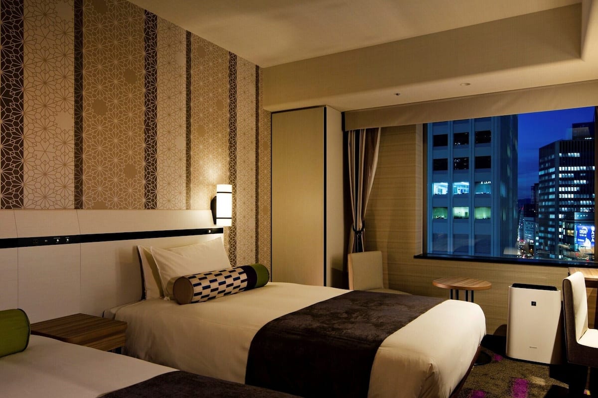 A hotel room with two beds, a desk, a chair, a window with a city view, and a patterned wall.