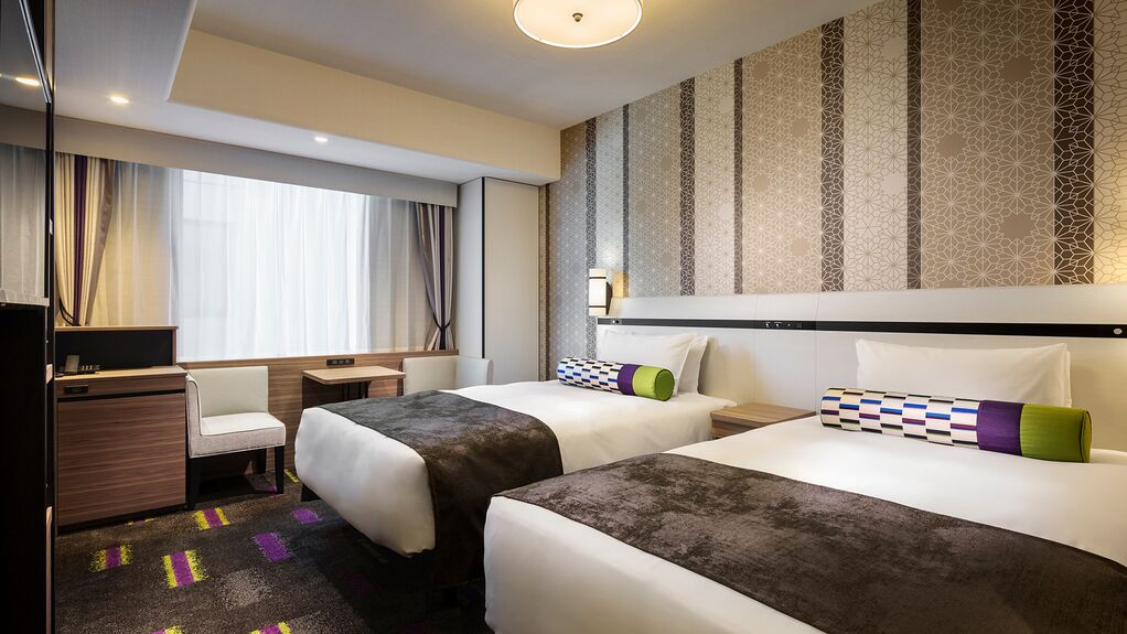 Habitación de hotel con dos camas, un escritorio, una silla y un ventanal con cortinas.