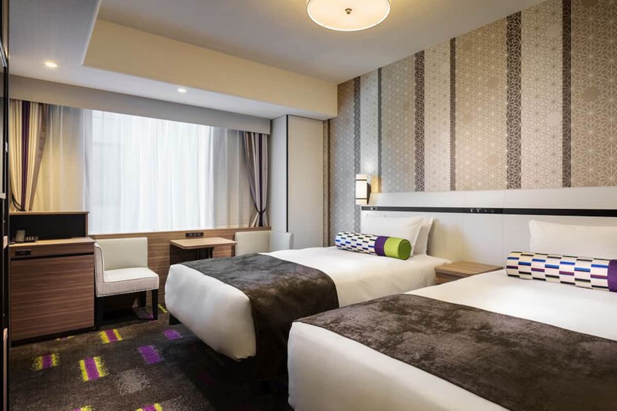 Habitación de hotel con dos camas, un escritorio, una silla y un ventanal con cortinas.