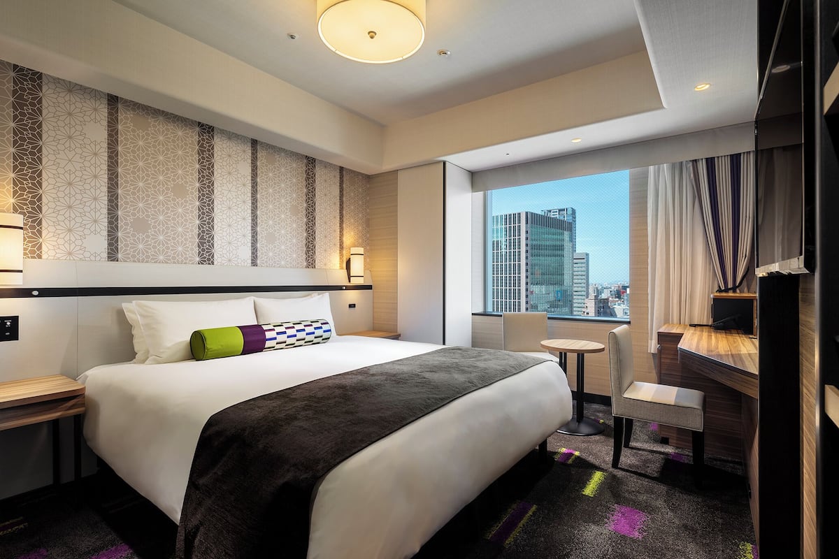 A modern hotel room with a large bed, a desk, a chair, and a view of the city through the window.