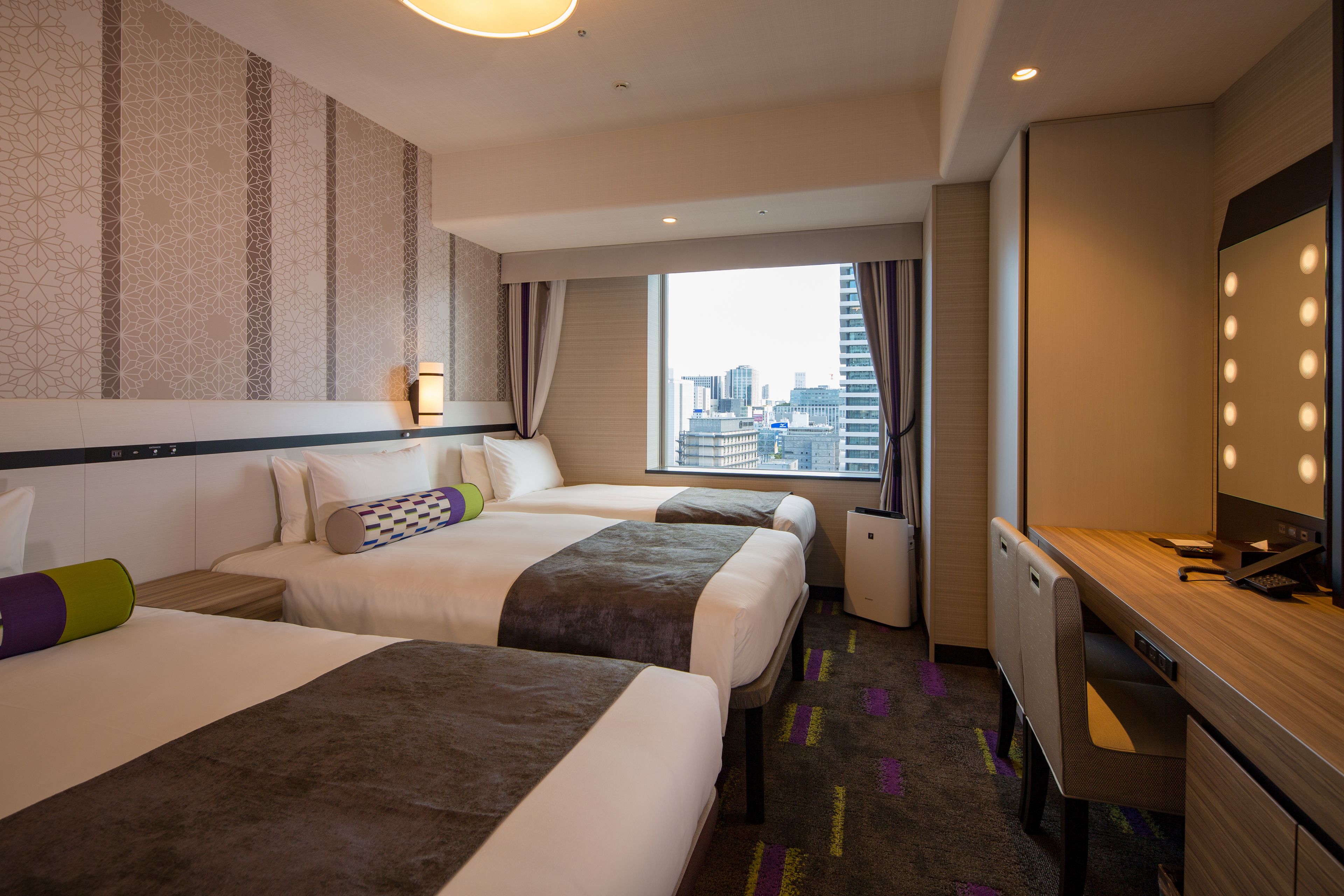 A hotel room with two beds, a desk, a mirror, and a window with a city view.