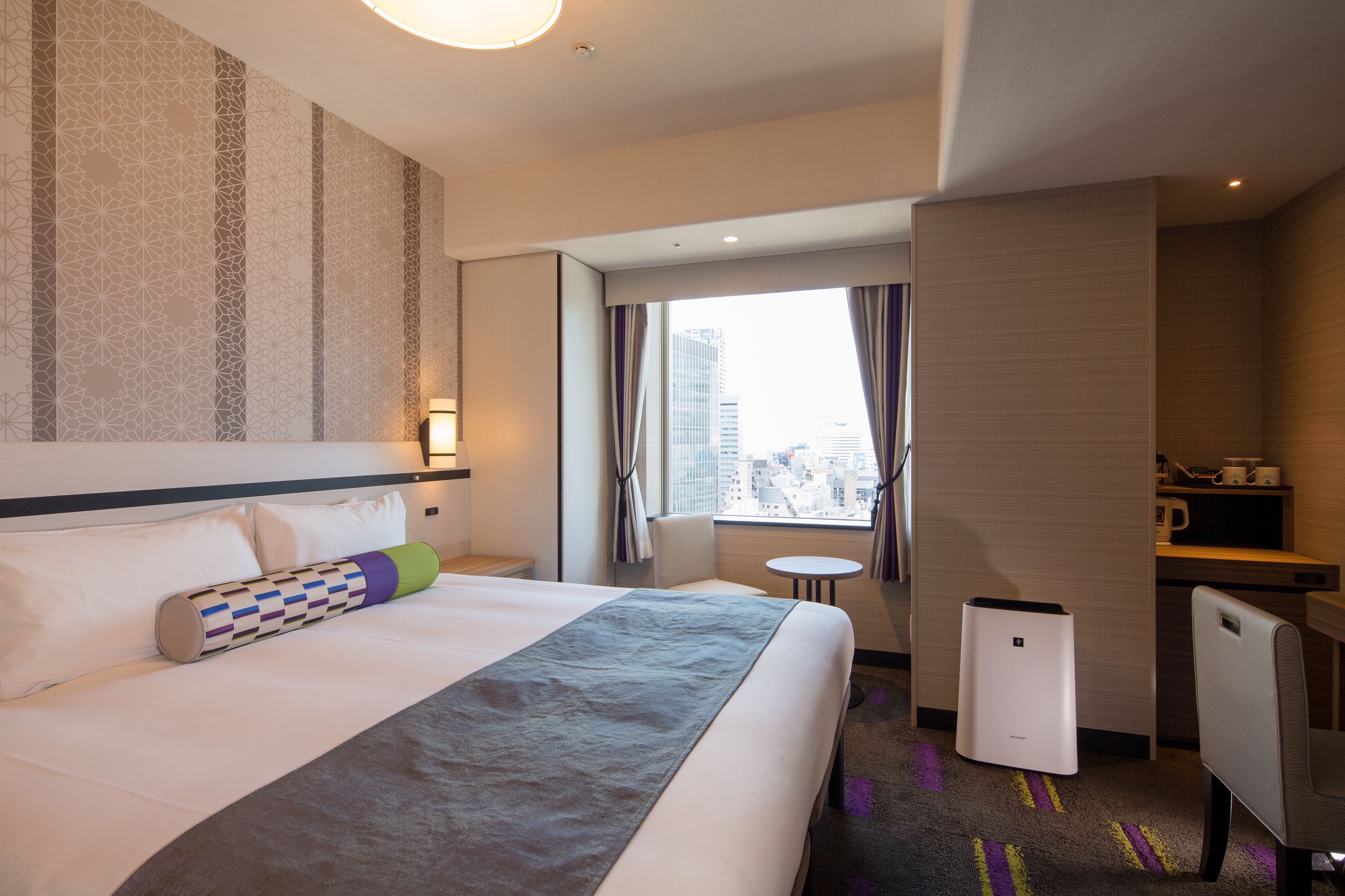 A hotel room with a large bed, a desk, a chair, a small table, and a view of the city through the window.