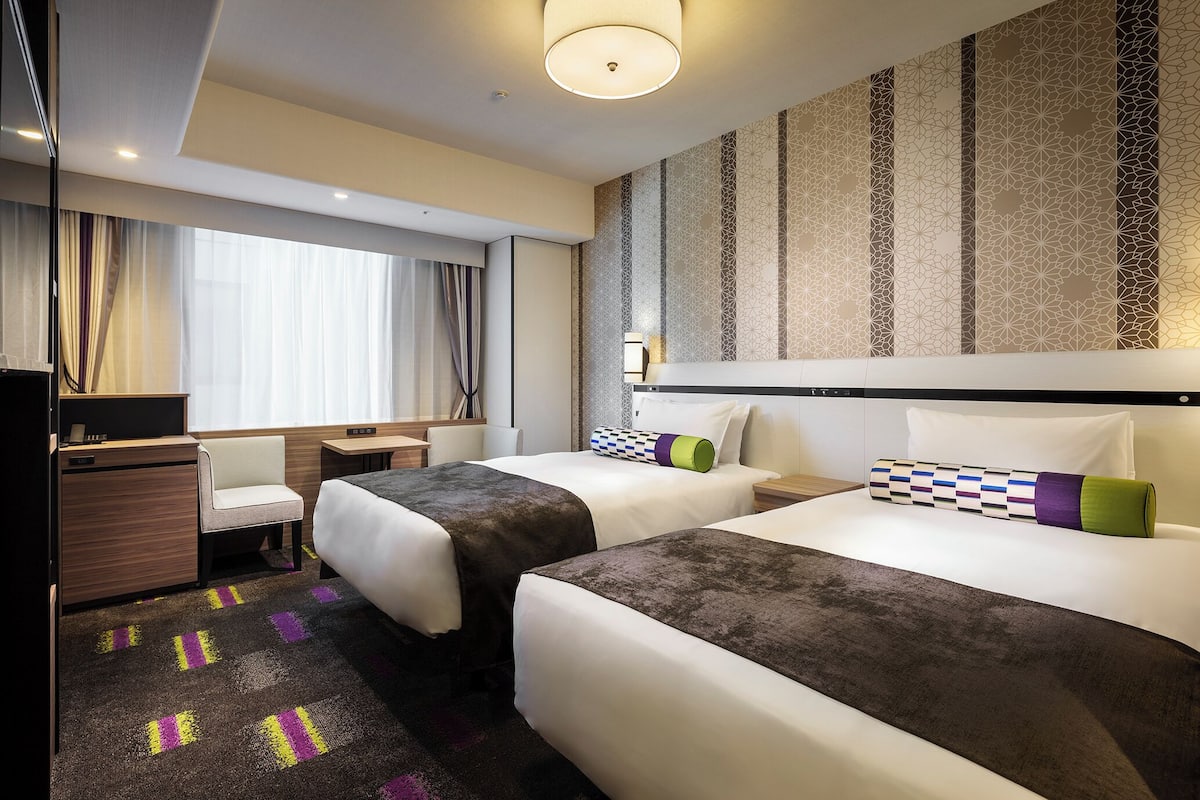 A hotel room with two beds, a desk, a chair, and a large window with curtains.