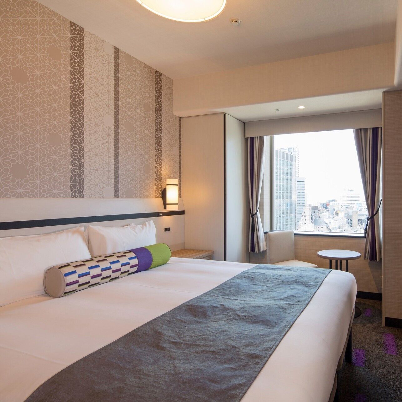 Habitación de hotel con una cama grande, una mesita, una silla, un ventanal con vistas a la ciudad y una pared con estampado.