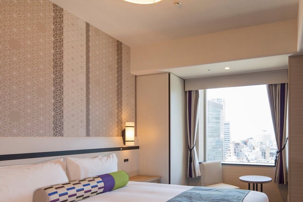 A hotel room with a large bed, a small table, a chair, a window with a city view, and a patterned wall.