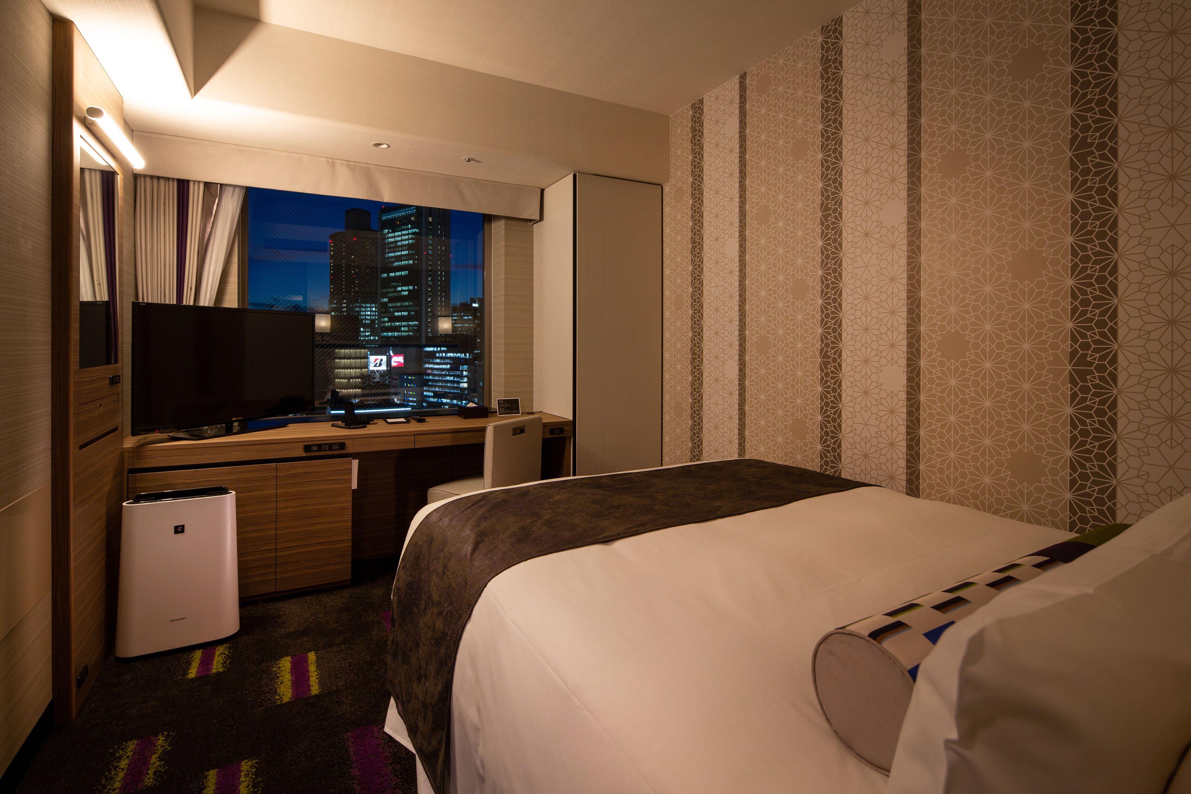 A hotel room with a bed, a desk with a computer, a television, and a view of a cityscape at night.