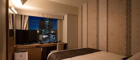 A hotel room with a bed, a desk with a computer, a television, and a view of a cityscape at night.