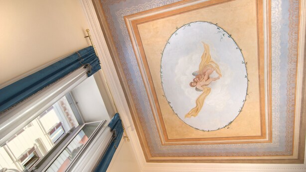A ceiling with a decorative painting of a cherub, a blue-framed window, and a patterned border. - Affitta Camere Via Veneto (Rome)