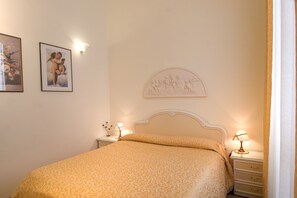 A neatly made bed with a patterned bedspread, a nightstand with a lamp, and wall art. - Affitta Camere Via Veneto (Rome)