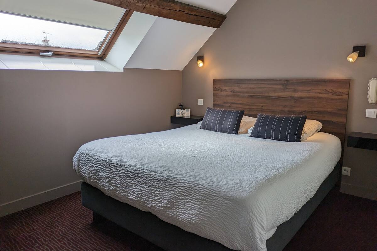 Une chambre avec un plafond en pente, un grand lit recouvert d’une couette blanche à texture, deux coussins à rayures, une tête de lit en bois et une petite table de chevet avec une lampe.