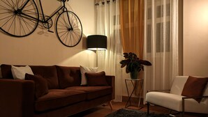 A living room with a brown sofa, a white armchair, a small table with a plant, a floor lamp, and a bicycle mounted on the wall. - Hôtel Le Saint Florent (Mont-pres-Chambord)