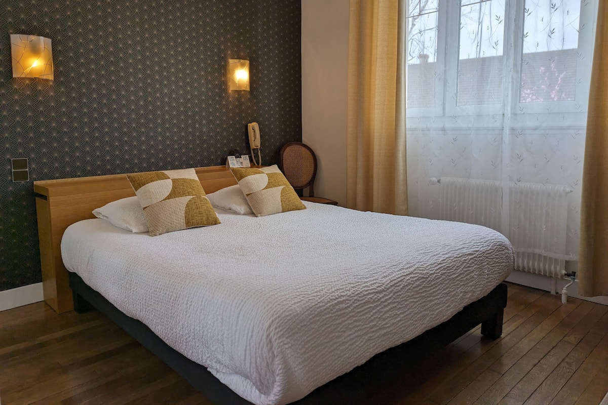Un lit bien fait, recouvert d’une couette blanche, avec une tête de lit en bois et plusieurs oreillers. Deux appliques murales et un téléphone sont fixés au mur. On aperçoit une fenêtre avec des rideaux légers, et un radiateur se trouve sous la fenêtre.