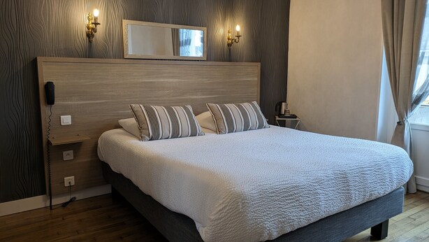 A modern bedroom with a wooden headboard, a large bed with white bedding, two bedside lamps, a nightstand with a coffee maker, and a mirror above the bed. - Hôtel Le Saint Florent (Mont-pres-Chambord)