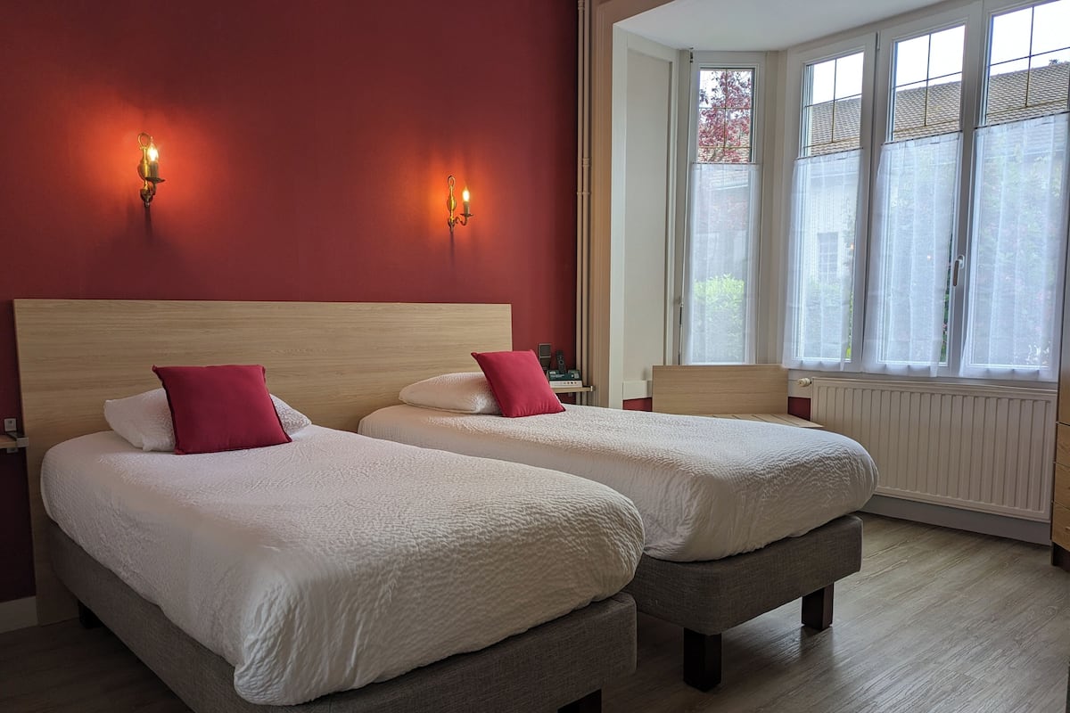 Une chambre d’hôtel avec deux lits, une tête de lit en bois, une fenêtre avec des rideaux légers et une table de chevet avec une lampe.