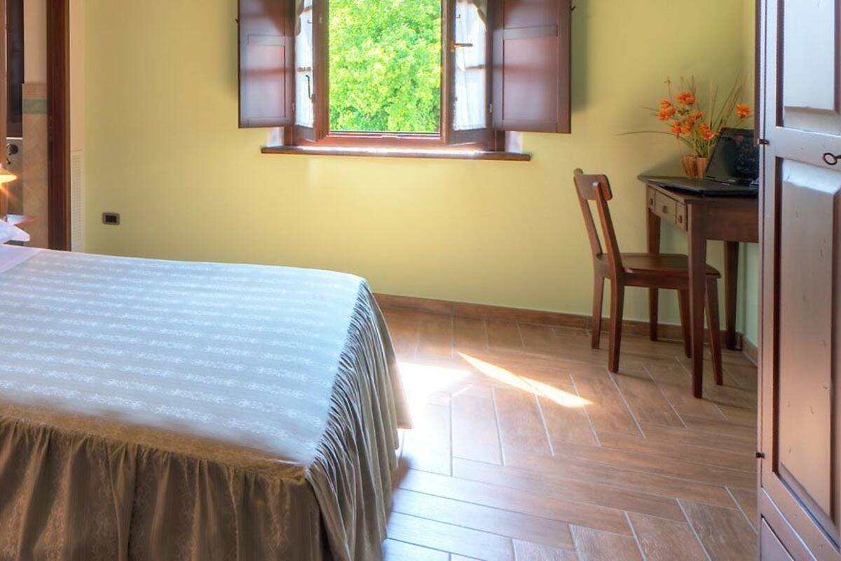 A bedroom with a bed, bedside lamps, a desk with a chair, and a window with a view of greenery.