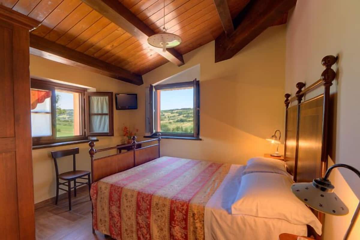 A bedroom with a wooden ceiling, a bed with a patterned bedspread, a chair, a desk, and a window with a view of the countryside.