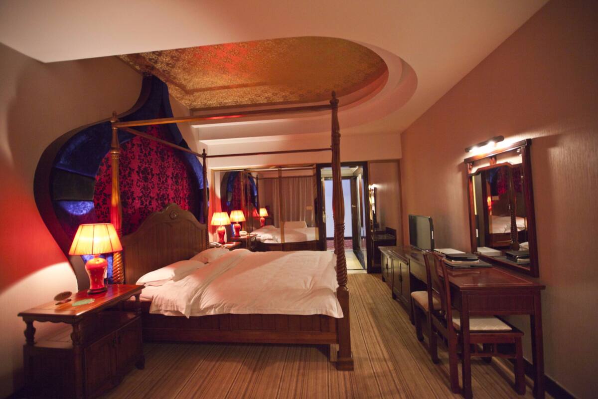 A four-poster bed with a canopy, a bedside table with lamps, a mirror, a desk with a chair, and a television.