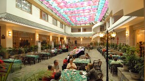 A spacious indoor dining area with multiple tables and chairs, surrounded by potted plants and traditional architectural elements. - YangShuo New Century Hotel (Guilin)