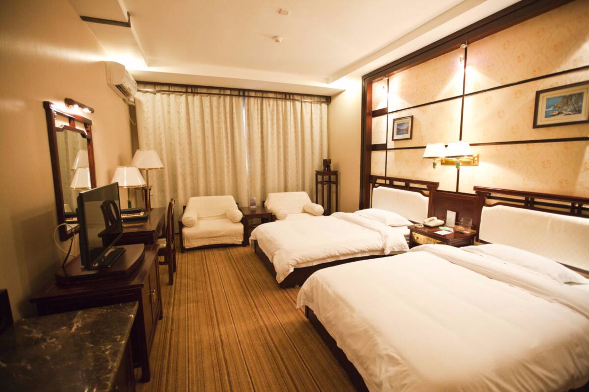 A hotel room with two beds, a desk with a computer, a chair, a bedside table with a lamp, and a large mirror.