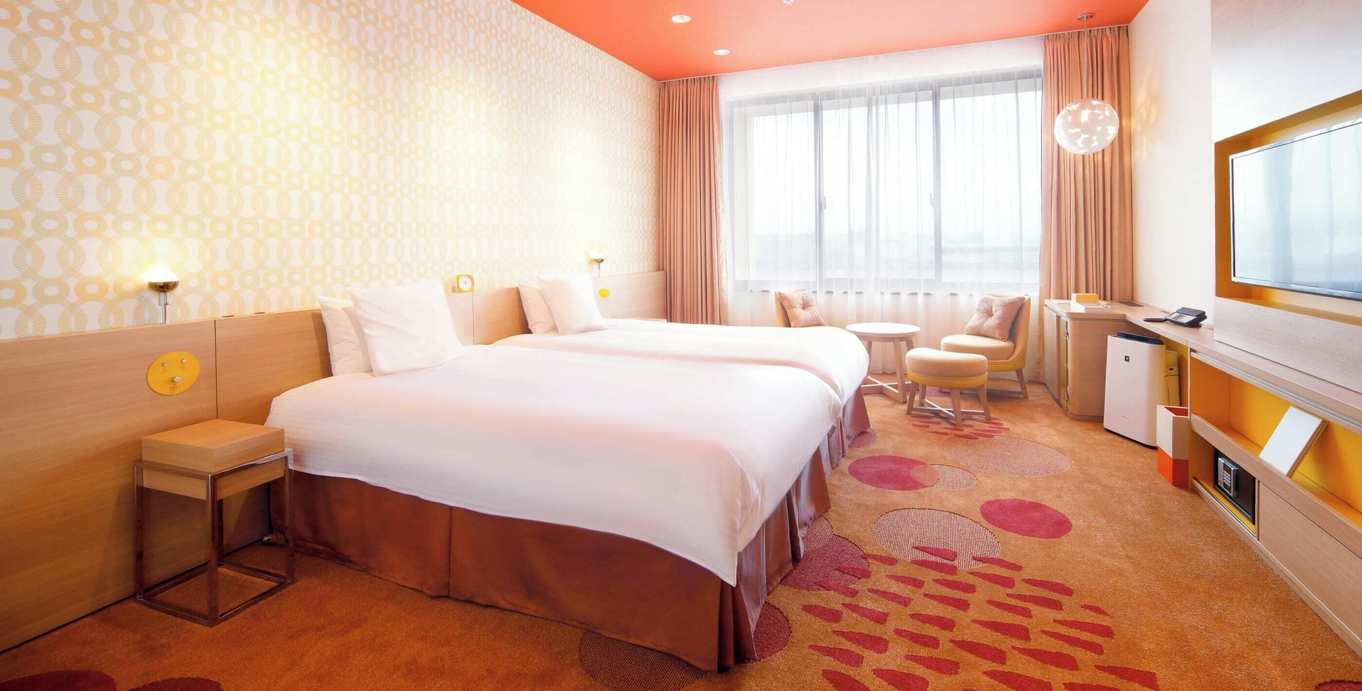 A hotel room with a large bed, bedside tables, a desk, a chair, a TV, and a window with curtains.