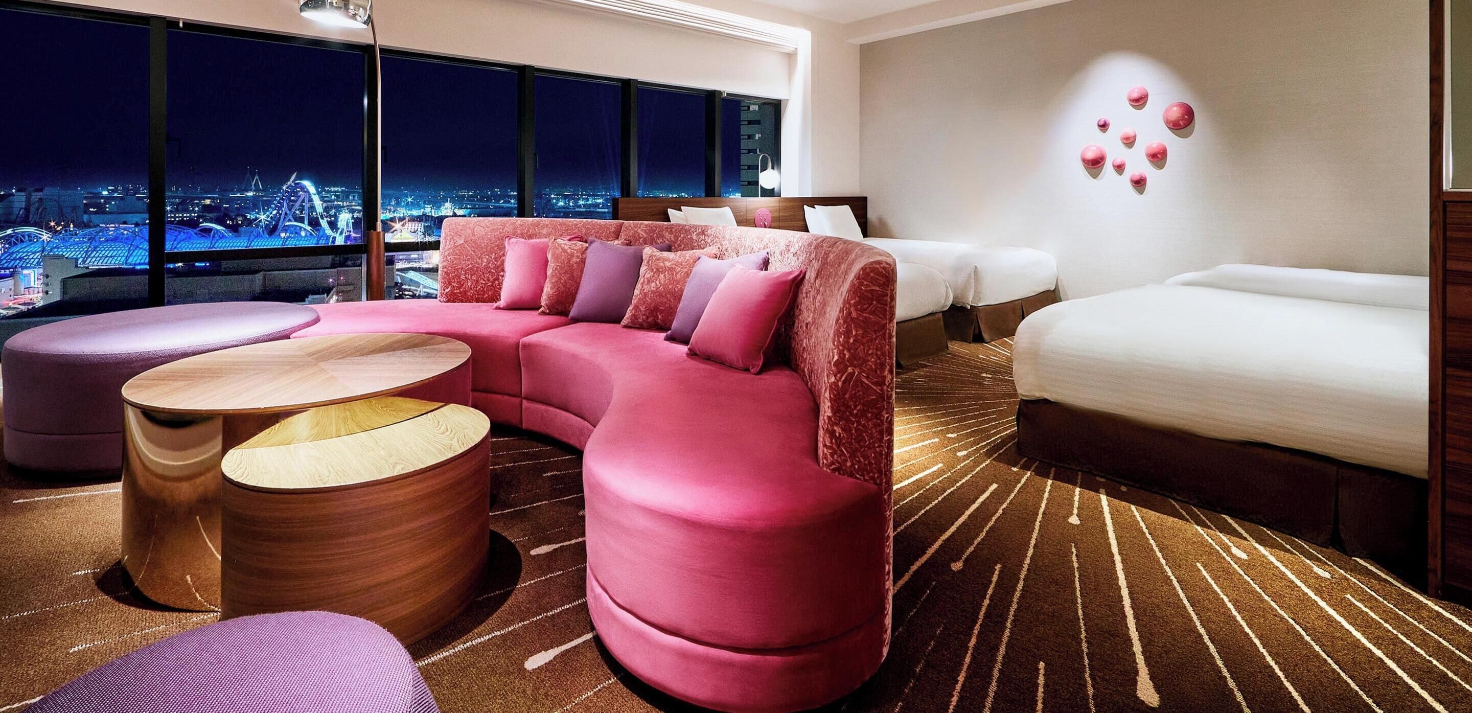 A modern hotel room with a large window, a pink sofa, a white bed, and a wooden coffee table.
