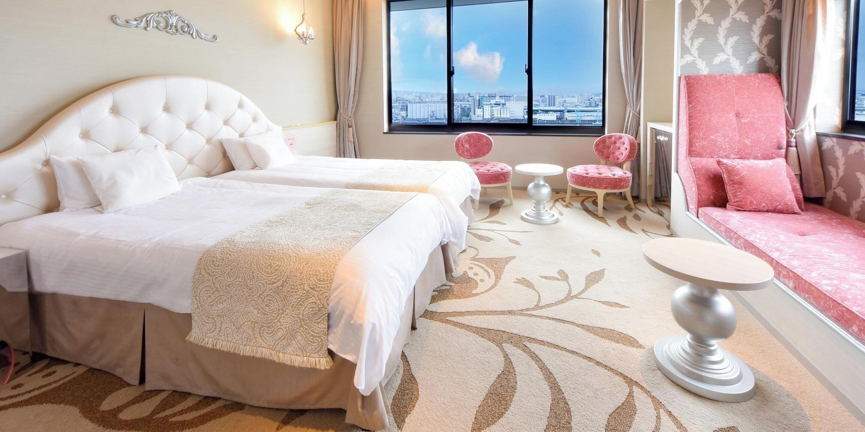 A hotel room with a large bed, two bedside tables, a seating area with a pink chair, and a view of the city through the window.