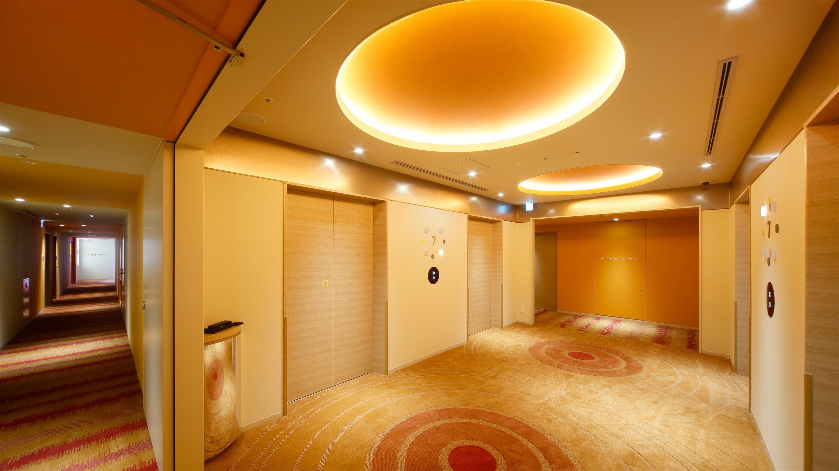 A hotel corridor with carpeted floors, wooden doors, and a ceiling with circular light fixtures.