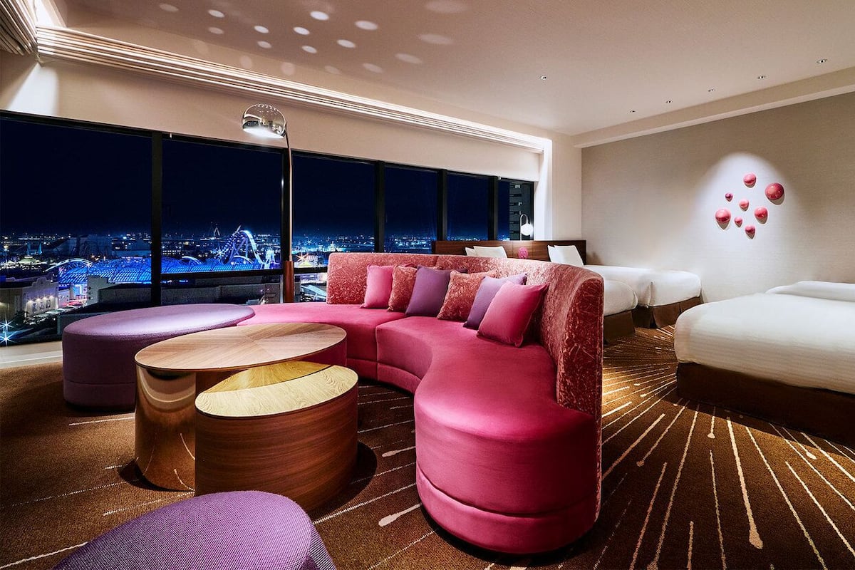 A modern hotel room with a curved pink sofa, a round wooden coffee table, a white bed, and a city view through large windows.