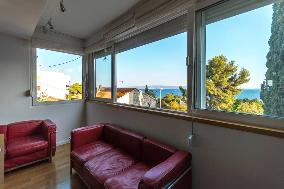 A room with a red leather sofa and a red armchair, large windows with a view of the sea and buildings.